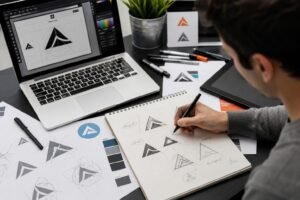 Graphic designer creating logo concepts in a sketchbook beside a laptop in a modern design studio